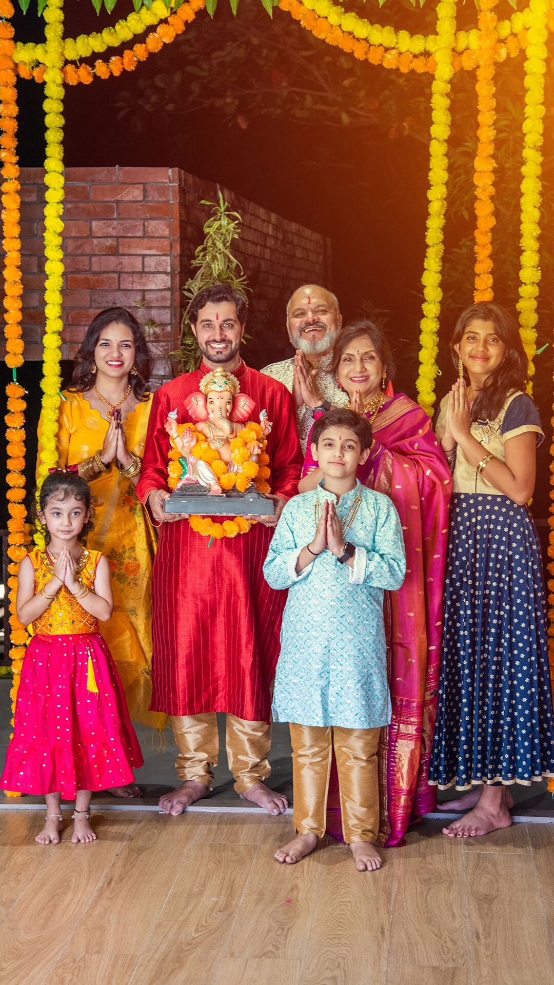 Indian family celebrating Ganesh Chaturthi at home, posing with Lord Ganesha idol, wearing traditional colorful outfits, surrounded by marigold flower decorations